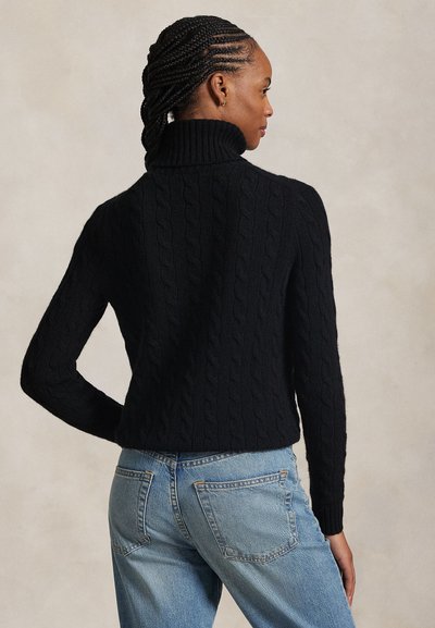 Donna con capelli intrecciati che indossa un maglione a collo alto nero a trecce e jeans blu, in piedi con una mano sui fianchi, rivolta di lato.