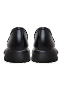 Paire de chaussures en cuir noir sans lacets avec des semelles en caoutchouc épaisses et crantées, vues de dos sur fond blanc.