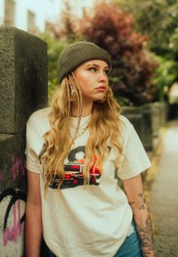 Light-colored t-shirt with pixel design, blue jeans, olive beanie, long blonde hair, gold chain, and large gold hoop earrings.