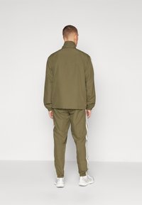 Tuta verde oliva con giacca con zip e pantaloni jogger. Presenta strisce laterali bianche e polsini elastici alle caviglie e alle maniche.