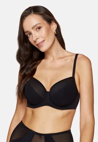 Czarny biustonosz z fiszbinami, z przezroczystymi siateczkowymi detalami, usztywnionymi miseczkami, regulowanymi ramiączkami i gładkim wykończeniem. Modelka w pozie z delikatnym uśmiechem.