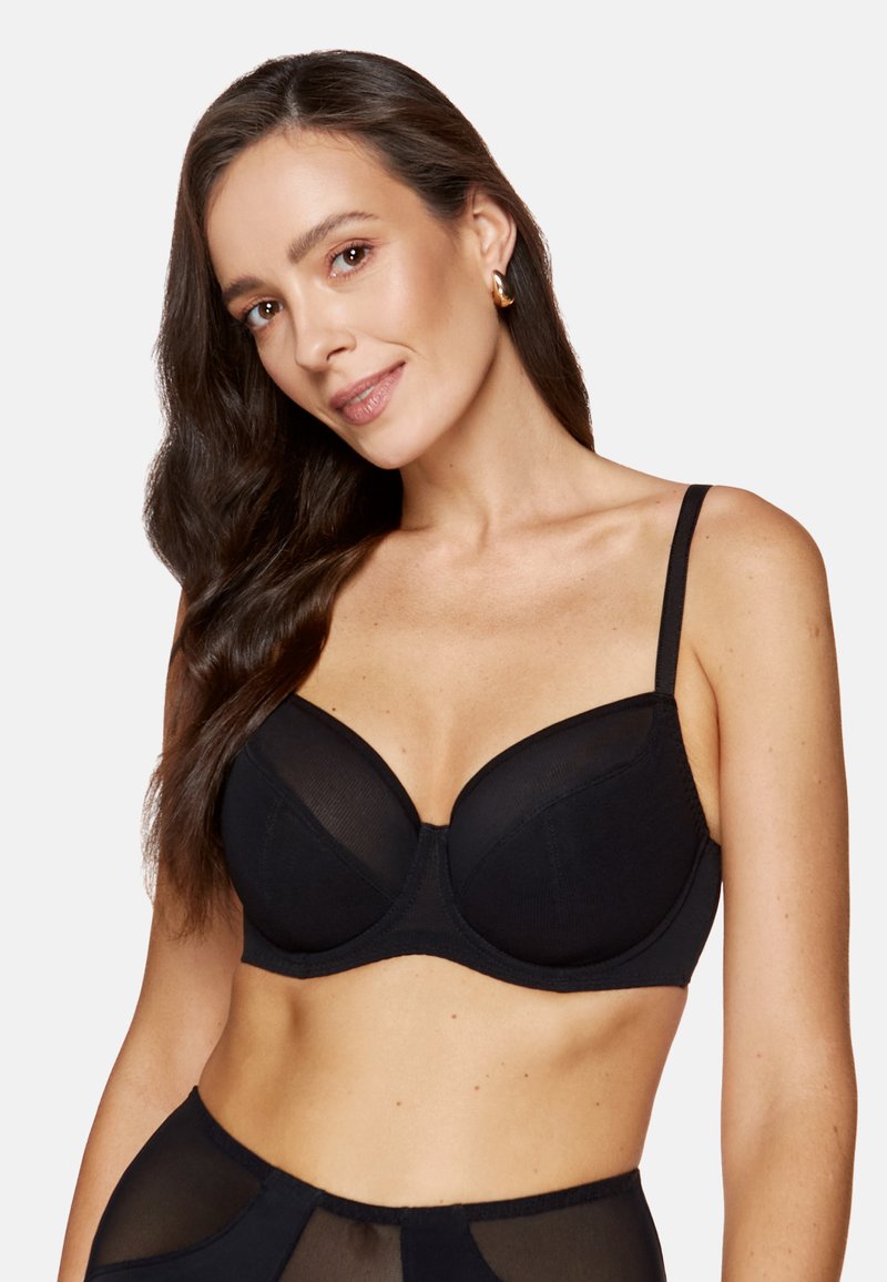 Czarny biustonosz z fiszbinami, z przezroczystymi siateczkowymi detalami, usztywnionymi miseczkami, regulowanymi ramiączkami i gładkim wykończeniem. Modelka w pozie z delikatnym uśmiechem.