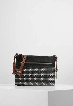Sac bandoulière à motif noir et blanc avec sangle réglable marron, poche avant zippée et petit porte-clés en or suspendu sur une surface blanche.