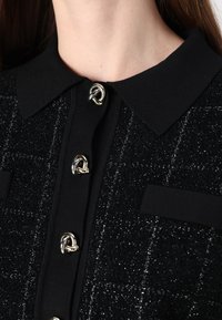 Blusa nera con texture, colletto e bottoni a nodi argentati lucidi. Presenta un delicato motivo a scacchi e due tasche sul petto.