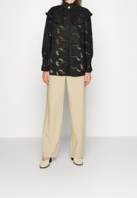 Haut en dentelle noire avec un col montant à volants et des manches longues, présentant un motif floral ajouré. Associé à un pantalon beige à jambes larges et des chaussures.