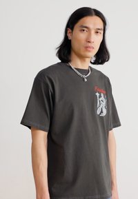 T-shirt en coton gris foncé avec un imprimé graphique sur le côté gauche de la poitrine, coupe décontractée et manches courtes avec des accents de couture visibles.