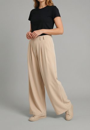 Donna che indossa una maglietta nera a maniche corte, pantaloni beige a vita alta e gamba larga, e sneakers beige, in piedi davanti a uno sfondo grigio.