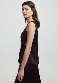 Sleeveless, deep burgundy top with a textured finish, featuring gold accents at the waist and a relaxed fit.