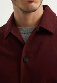 Veste bourgogne avec une fermeture à boutons, dotée d'un col classique et d'un tissu texturé. Portée par-dessus un pull en tricot gris.