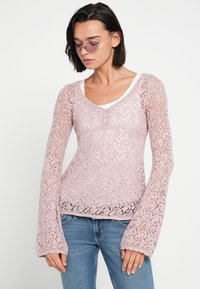 Απαλό ροζ πλεκτό πουλόβερ με δαντελένιο σχέδιο, μακριά φαρδιά μανίκια και στρογγυλή λαιμόκοψη· φορεμένο πάνω από λευκή μπλούζα.