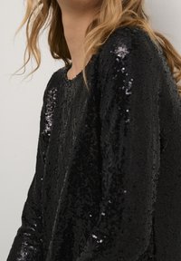 Närbild av en person som bär en långärmad svart paljettopp, som visar textur och glans, med blont hår delvis synligt.