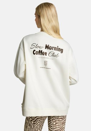 Femme blonde de dos portant un sweat blanc avec le texte "Slow Morning Coffee Club" et un pantalon à imprimé zèbre sur un fond uni.