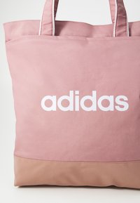 Roza torba na rame s velikim bijelim "adidas" logotipom, izrađena od glatke tkanine s kontrastnim bež donjim dijelom i čvrstom ručkom.
