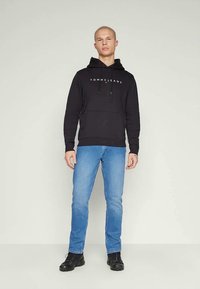 Μαύρη κουκούλα φούτερ με το λογότυπο "TOMMY JEANS", τσέπη καγκουρό, συνδυασμένη με μπλε τζιν και μαύρα υποδήματα. Απλός, άνετος σχεδιασμός.