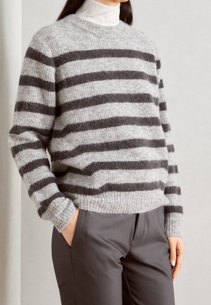 Personne portant un pull douillet rayé gris et gris foncé sur un col roulé blanc, avec un pantalon gris et une main dans la poche.