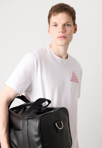 Black leather bag with a rectangular shape, featuring a top handle and a silver ring accent, held against a white t-shirt with red logo print.