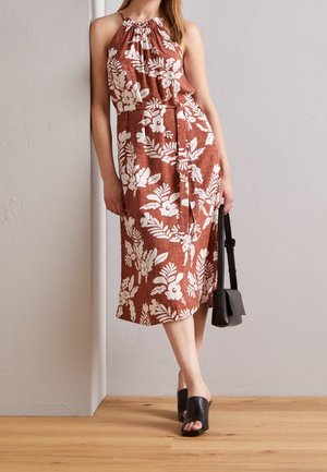 Frau trägt ein ärmelloses braunes Kleid mit weißem Blumenmuster, hält eine schwarze Handtasche und trägt schwarze offene Mule mit Absatz.