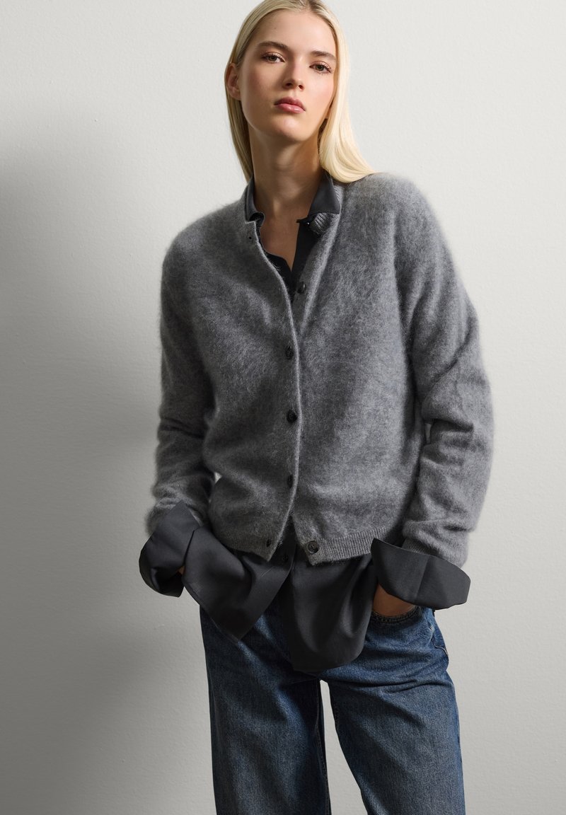 Grauer, gestrickter Cardigan aus weichem, flauschigem Material, mit Knopfleiste, runder Halslinie und geripptem Saum, über einer dunklen Bluse getragen.
