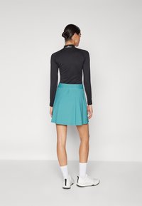 Svart langermet topp kombinert med en plisséformat turkise skjørt. Modellen har på seg hvite joggesko og hvite sokker, noe som viser en sporty stil.