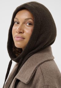 Palton din lână maro cu o glugă mare și întunecată. Textură netedă, guler supradimensionat și detalii cu șnururi. Modelul are părul creț ieșind de sub glugă.