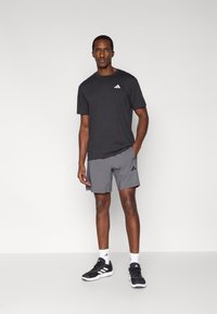 Ανδρική αθλητική στολή: μαύρο μπλουζάκι με κοντά μανίκια, γκρι σορτς, μαύρα αθλητικά παπούτσια με λευκές λεπτομέρειες και λευκές αθλητικές κάλτσες. Λογότυπο εμφανές στο μπλουζάκι και τα σορτς.