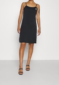 Vestido negro sin mangas de tela suave, con un diseño sencillo y un pequeño logotipo en el pecho. Tirantes delgados y corte a la altura de la rodilla.