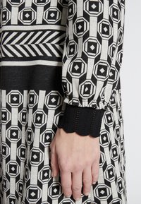 Abito a righe bianche e nere che mette in evidenza forme geometriche, con un polsino ondulato nero testurizzato alle maniche e una finitura in tessuto liscia.
