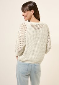Weißer Strickpullover mit offenem Maschenmuster, rundem Ausschnitt und überschnittenen Schultern. Kombiniert mit hellblauen Jeans mit Gesäßtaschen.