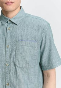 Camisa de manga corta, de color azul claro, hecha de tela texturizada. Presenta un cierre de botones en la parte delantera y un bolsillo en el pecho con un borde azul en contraste.