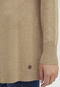 Beige kötött pulóver bordás mandzsettával és aljjal, kis fém logóval a alsó részen. Sima textúrájú, laza fazonú.