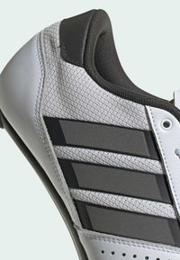 Detailní záběr na bílé sportovní boty s černým polstrovaným límcem a třemi širokými tmavými pruhy na síťovaném bočním panelu.