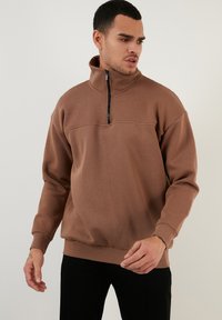 Sudadera de color marrón con cuello alto, cremallera de cuarto y ribetes en el bajo y las muñecas. Tela suave y corte relajado para mayor comodidad.