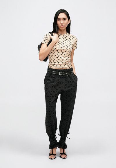 Bézs színű, testhezálló póló fekete félhold mintával; fekete nadrág tónusos mintával és állítható bokapántokkal; fekete pántos szandálok.