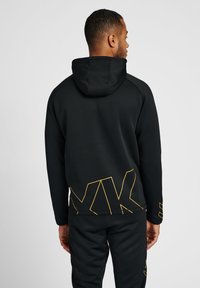 Man met de rug naar de kijker, gekleed in een zwarte hoodie en broek met een geel geometrisch lijnontwerp op de onderrug en mouwen, tegen een effen achtergrond.