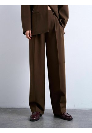 Person iført løse brune bukser og matchende blazer, suppleret med mørkerøde loafers, stående på gråt gulv mod hvid væg.