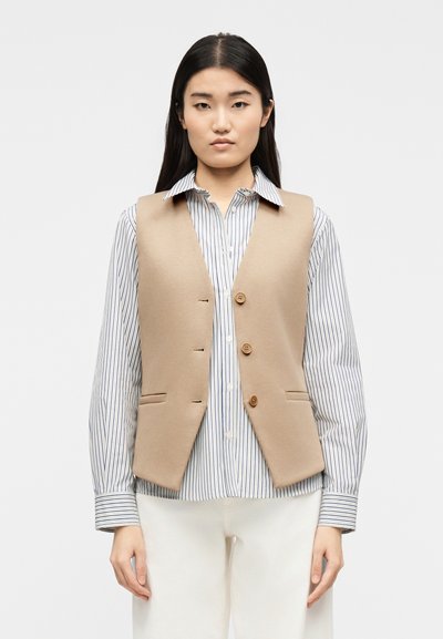 Joven mujer con un chaleco beige abotonado sobre una camisa de manga larga a rayas y pantalones blancos, de pie frente a un fondo liso.