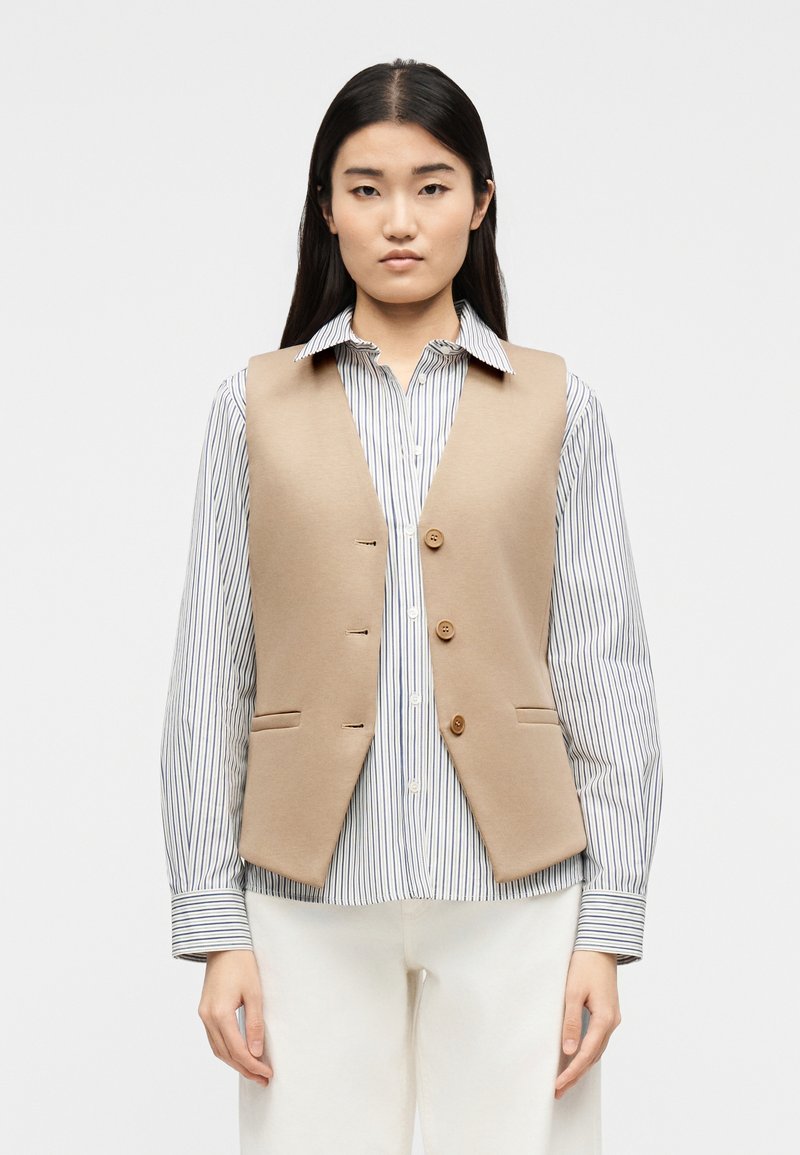 Νεαρή γυναίκα που φοράει ένα μπεζ γιλέκο με κουμ πιά πάνω από μια ριγέ μπλούζα μακρυμάνικη και άσπρο παντελόνι, στέκεται μπροστά σε απλό φόντο.