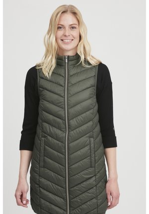 Gilet imbottito verde e isolato con motivo a chevron, colletto alto e cerniera frontale. Indossato sopra un top a maniche lunghe nero, presenta tasche laterali.