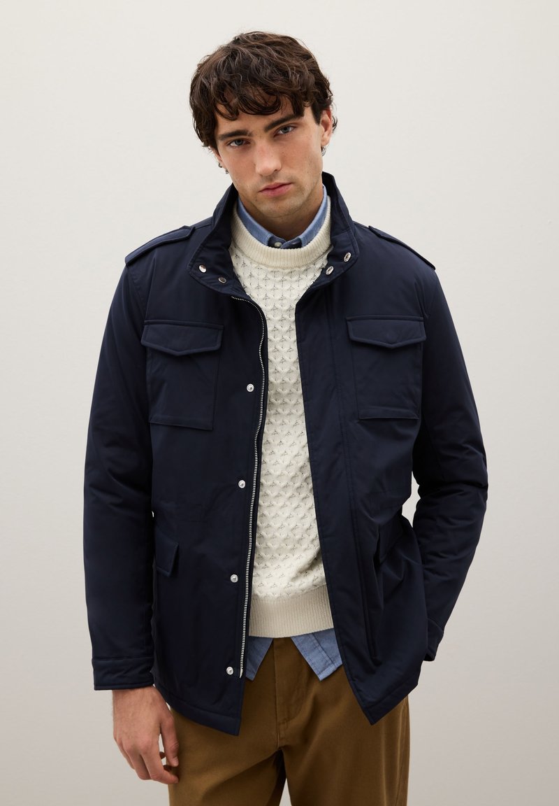 Giacca blu navy con due tasche frontali, chiusura con zip e collo alto, indossata sopra un maglione crema lavorato a maglia e una camicia blu.