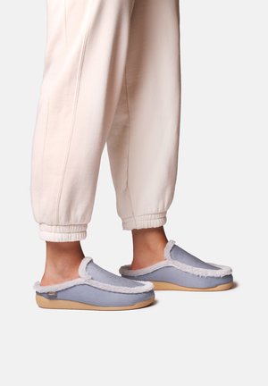 Personne portant des chaussons bleu clair à enfiler avec bordure en fourrure blanche et semelles beige, associés à un pantalon ample couleur crème.