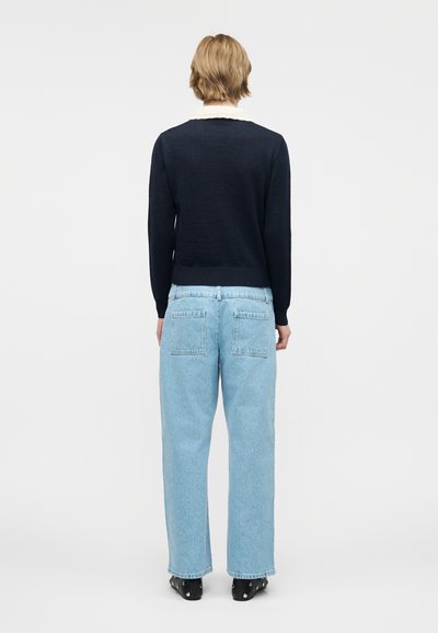 Άτομο με κοντά ξανθά μαλλιά, φορώντας μαύρο πουλόβερ με μακριά μανίκια, ανοιχτό μπλε τζιν παντελόνι φαρδύ πόδι και μαύρες μπότες, στέκεται με την πλάτη.