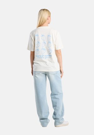 Camiseta color crema con un patrón gráfico de fresas azules en la parte posterior, texto "Sabor de Verano" y "Garden Grove," combinada con jeans de color azul claro.