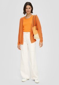 Leichter orangefarbener Cardigan über einem orangefarbenen Top, kombiniert mit weißen weit geschnittenen Hosen und weißen Sneakers. Das Modell hält eine geflochtene Handtasche.