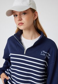 Granatowy sweter z zamkiem błyskawicznym w poziome białe paski, z ribbowymi mankietami i haftem "Los Angeles", noszony na białej koszulce.
