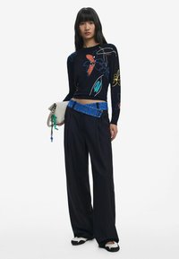 Schwarzes Langarmoberteil mit buntem floralem Stickerei, kombiniert mit weiten schwarzen Hosen, die einen blauen Jeansgürtel und eine passende Clutch-Handtasche haben.