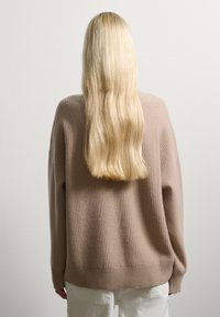 Beige gestrickter Pullover mit gerippter Textur. Hat einen lockeren Schnitt und lange Ärmel, kombiniert mit weißen Hosen. Die Haare fallen den Rücken hinunter.