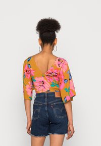 Top corto floral con flores vibrantes en rosa y azul sobre un fondo mostaza, con mangas anchas y diseño de espalda en V, combinado con pantalones cortos de mezclilla.