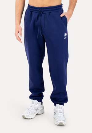 Uomo che indossa pantaloni della tuta blu navy con coulisse e un piccolo logo sulla coscia sinistra, con scarpe da ginnastica bianche, in piedi contro uno sfondo semplice.