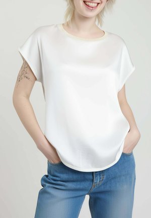 Persona sorridente con capelli biondi corti che indossa una maglietta bianca a maniche corte e jeans blu con le mani nelle tasche, mostrando un tatuaggio sul braccio e un piercing al labbro.