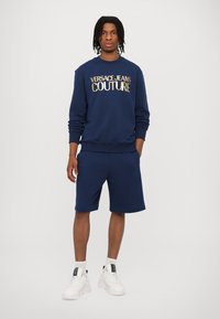 Temno modra bombažna majica s kapuco z napisi "VERSACE JEANS COUTURE" v zlatih črkah, v kombinaciji s usklajenimi kratkimi hlačami in belimi športnimi copati. Gladka struktura, udoben kroj.
